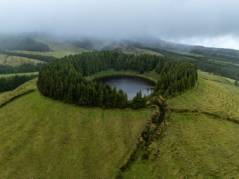 Aerial view of Lagoa Pau Pique surrounded by a dense ring of evergreen trees on a lush green volcanic crater, Capelas, Azores, Portugal.