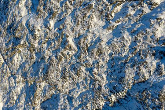 Aerial view of a steep, snow-covered rocky mountain slope with rugged textures and deep shadows under bright daylight in Valldal, More og Romsdal, Norway.