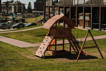 Wooden playground with climbing wall, slide and swings stands on a grassy lawn in Kopaonik. Resort hotels and leisure facilities appear in the background © Ekaterina