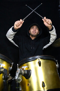 Young male drummer in a black and white hoodie performing on a gold drum set against a black background. Excited musician holding crossed drumsticks in the air with passion and energy during a live pe