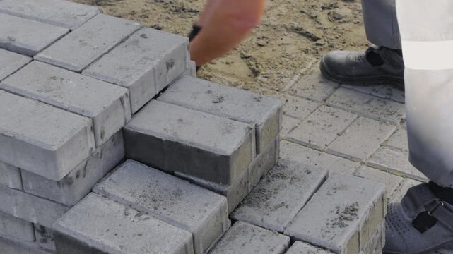 Construction worker installs gray concrete pavers on a sand base. Close-up of manual pavement work for landscaping and road improvement.