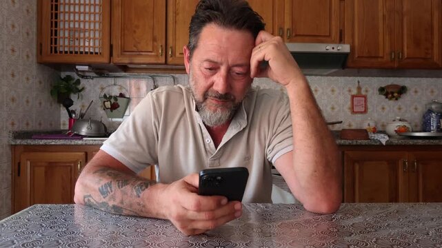 A middle-aged man with a beard sits at his kitchen table, looking sadly at his smartphone, seemingly distressed or worried. He rubs his eyes and forehead in a gesture of fatigue and concern, reflectin