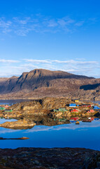 scenic view of the coastal town of Sisimiut, Greenland, featuring brightly painted wooden houses nestled against rugged, rocky terrain and mirrored in the calm fjord waters © Janos