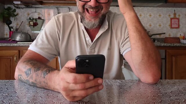 A smiling man with a beard and glasses sits at a kitchen table, looking happily at his smartphone. He seems amused or engaged with content on his phone, showing a pleasant and relaxed demeanor.