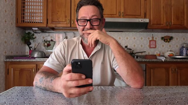 A cheerful middle-aged man in a home kitchen is happily engaged with his mobile phone, smiling and laughing while looking at the screen. He appears to be enjoying content or a conversation.