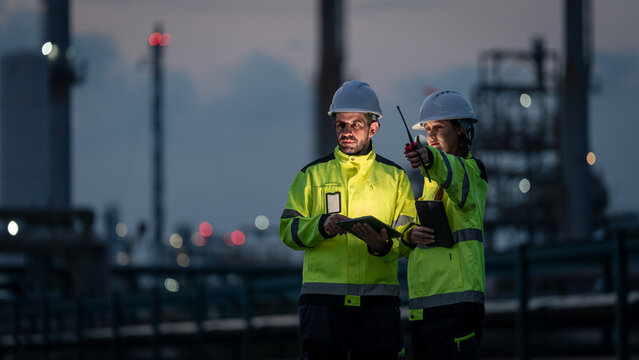 Refinery Engineering Team Reviewing Digital Tablets and Pointing toward Industrial Equipment while Wearing Reflective Jackets and Hard Hats at a Petrochemical Plant after Sunset