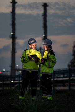 Refinery Engineering Team Reviewing a Digital Tablet and Communicating by Handheld Radio while Wearing Reflective Safety Jackets and Hard Hats at a Petrochemical Plant after Sunset