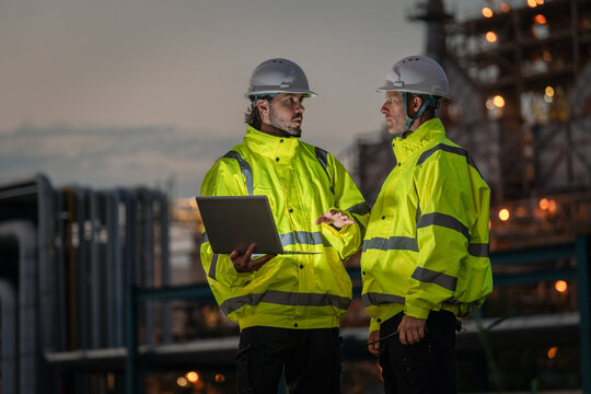 Refinery Engineers Discussing Field Operations with an Open Laptop while Wearing Reflective Safety Jackets and Hard Hats beside Illuminated Petrochemical Equipment after Sunset