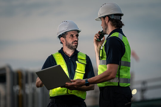 Refinery Engineers Coordinating Field Operations with a Laptop Computer and Handheld Radio while Wearing Reflective Safety Vests and Hard Hats at an Industrial Site during Evening Work