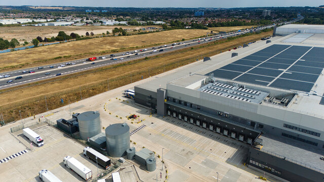 Aerial view of a large supermarket distribution centre with loading bays, parked trucks, and solar panels on the roof, located next to a busy highway in Luton, England, United Kingdom.