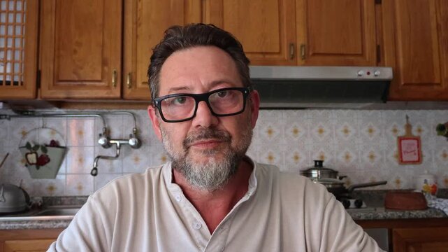 A middle-aged man with glasses and a beard, in a home kitchen, momentarily sticks out his tongue, then looks directly into the camera with a neutral expression.