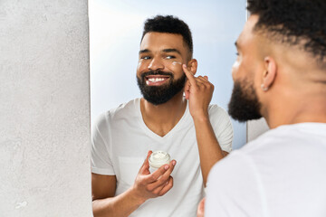 A young, handsome Black man smiles in front of a mirror while applying face cream.