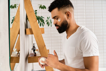 A young man applies moisturizer to his face while standing in his stylish home bathroom.