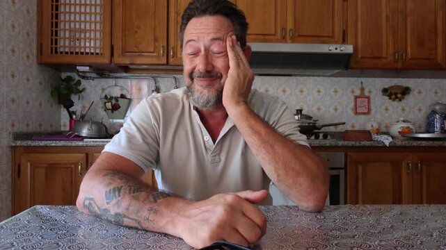 A middle-aged man with a beard and tattoos sits thoughtfully at a kitchen table, experiencing moments of stress and reflection. He covers his eyes, showing weariness and possibly deep thought or worry