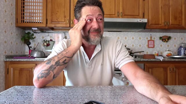 A middle-aged man in a kitchen rubs his eyes, showing deep stress, tiredness, and worry. His pensive expression suggests he is grappling with a problem or a severe headache.