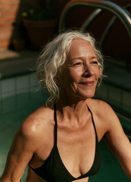 Portrait of an elderly woman with white curly hair. she is wearing a black bikini and is standing in front of a swimming pool. the woman is smiling and looking directly at the camera.