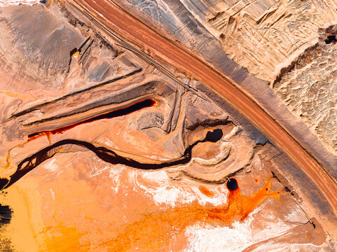 Aerial view of an open-pit mine with vibrant red earth, industrial mining infrastructure, and a dark tailings pond near a dirt road in Pinjarra, Western Australia, Australia.