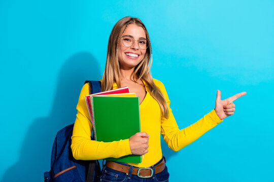 Smiling student woman with backpack holding colorful notebooks and pointing to the side against a bright blue background