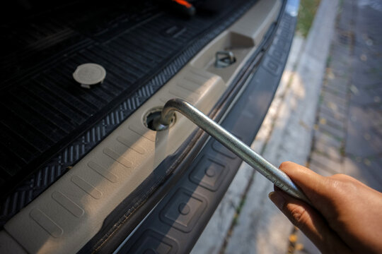Hand using a metal crank tool to access a vehicle's spare tire mechanism, illustrating car maintenance, tire replacement, roadside repair, and automotive emergency preparedness.