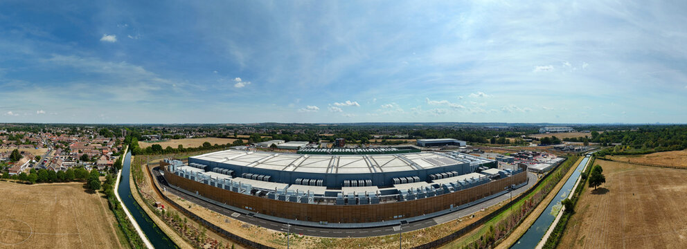 Aerial view of a large hyperscale data center featuring numerous air handling units and chillers on the roof, situated near a canal and fields in Hereford, England, United Kingdom.