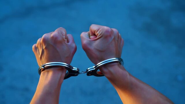 Handcuffed clenched fists held aloft against blue background with linked metal restraints