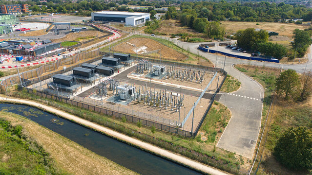 Aerial view of an electrical substation with high voltage transformers and equipment, surrounded by a security fence, near a canal in Waltham Cross, England, United Kingdom.