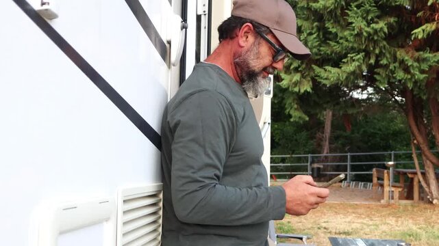 A happy bearded man in glasses and a cap leans against a camper van, smiling while using his smartphone outdoors. He enjoys a moment of connection and relaxation on his travel adventure.