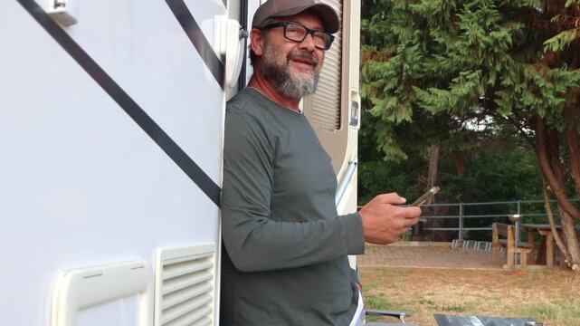 A man wearing a cap and glasses with a beard leans against a campervan, casually using his smartphone. He looks up and smiles, then returns to his device, enjoying a relaxed moment outdoors.