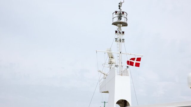 Danish flag waves from ship mast beneath cloudy sky, surrounded by navigation lights and maritime equipment. Close view captures wind, national symbol, vessel structure and seafaring atmosphere
