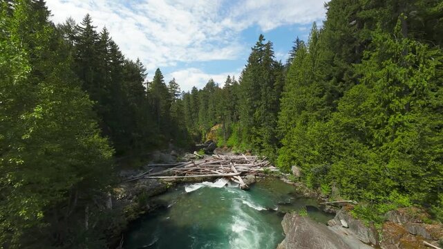A natural forward moving aerial camera glides over churning turquoise white water rapids and large boulders toward a rustic log jam under a patchy blue sky on a summer day.
