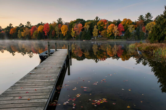 A wooden dock extends into a misty lake with a small boat moored beside it autumn trees with vibrant colorful leaves reflect on the calm water as a great blue heron stands by the shore