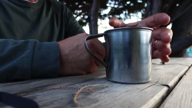 A person's hands rest contemplatively on a rustic wooden picnic table next to a metal mug, suggesting a moment of quiet reflection or a peaceful outdoor break amidst nature.