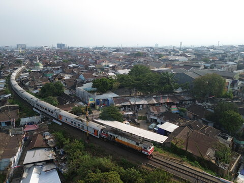 Aerial view of a passenger train traveling through the dense residential area with traditional rooftops and surrounding greenery in Blimbing, East Java, Indonesia.