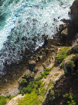 Aerial view of Baron Beach featuring rugged cliffs, lush green vegetation, and white sea foam crashing against dark rocks in Yogyakarta, Special Region of Yogyakarta, Indonesia.