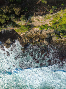 Aerial view of Baron Beach with crashing waves hitting the rocky shoreline, lush green vegetation on the cliffs, and clear blue water in Yogyakarta, Special Region of Yogyakarta, Indonesia.