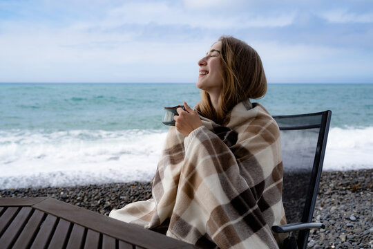 Happy woman wrapped in a warm blanket enjoying a hot drink by the sea on a cool and cloudy day. A cozy way of life and a concept of relaxing alone.