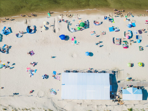 Aerial view of a sandy beach with colorful umbrellas, people relaxing on the sand, and a striped roof of a beach bar in Krynica Morska, Pomeranian Voivodeshi, Poland.