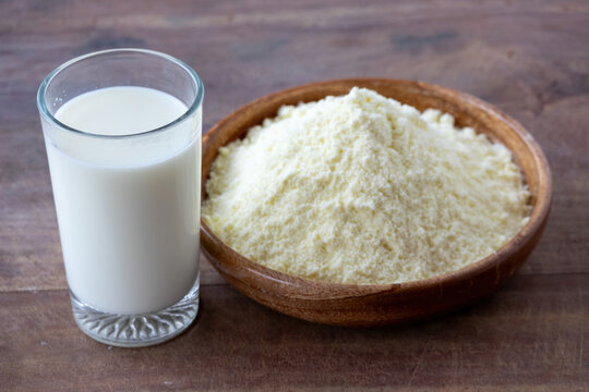 Fresh liquid milk filled in transparent glass served beside dry powdered milk in wooden bowl. Natural dairy nutrition product on wooden table background.
