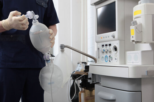 An anesthesiologist in blue uniform and gloves squeezes an Ambu breathing bag against background of an anesthetic device in clinic
