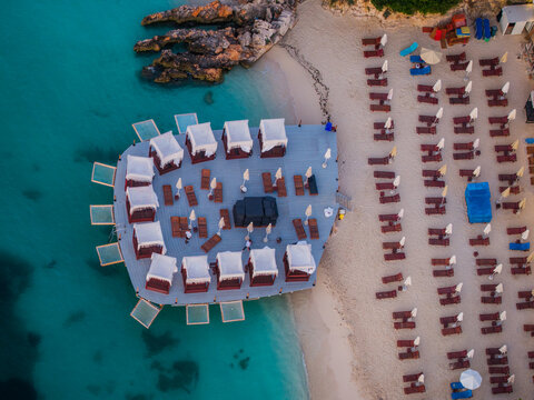 Aerial view of a beach club with white cabanas on a wooden deck over turquoise water, next to a sandy beach with rows of sun loungers and umbrellas in Ksamil, Vlore County, Albania.