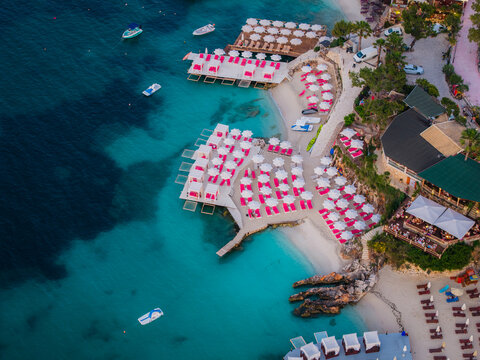 Aerial view of a beach resort in Ksamil with rows of pink sun loungers on wooden decks and white sand, surrounded by turquoise water and rocky coastline in Ksamil, Vlore County, Albania.