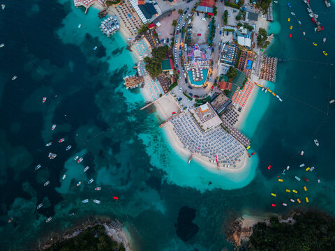 Aerial view of the turquoise coastal waters and sandy beaches of Ksamil featuring organized beach clubs with rows of umbrellas and small boats in Ksamil, Vlore County, Albania.