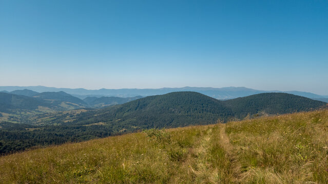 High-angle panoramic view of rolling mountain ridges, sloping grassy hillside, and expansive clear blue sky on a sunny day.