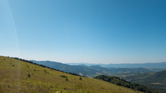 High-angle panoramic view of rolling mountain ridges, sloping grassy hillside, and expansive clear blue sky on a sunny day.