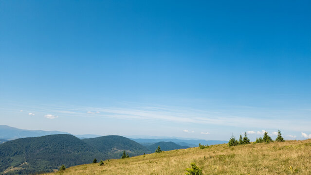 Panoramic landscape photograph of vast rolling green hills and distant mountain ranges under a wide, clear blue sky with light summer clouds. Concept for nature travel, wilderness hiking, and scenic g