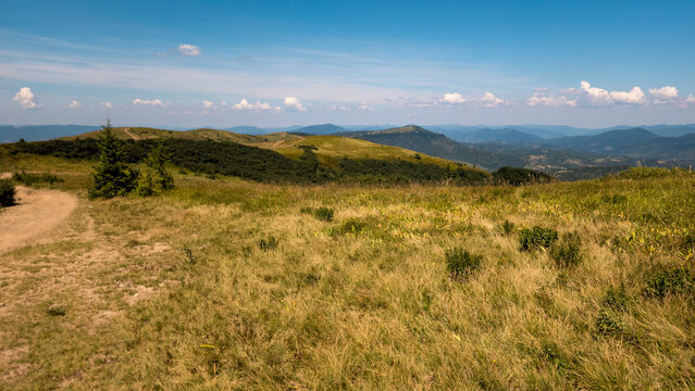 Panoramic landscape photograph of vast rolling green hills and distant mountain ranges under a wide, clear blue sky with light summer clouds. Concept for nature travel, wilderness hiking, and scenic g