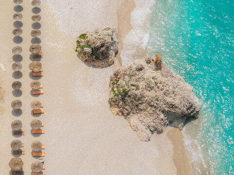 Aerial view of Kroreza Beach featuring turquoise water, large rocks on the sand, and rows of straw umbrellas with sun loungers in Sarande, Vlore County, Albania.