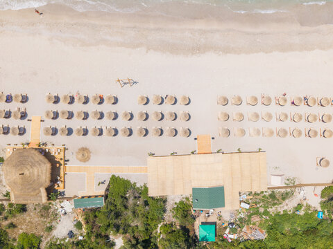 Aerial view of Kroreza Beach featuring rows of straw umbrellas on white sand, wooden walkways, and a beach bar surrounded by lush greenery in Sarande, Vlore County, Albania.