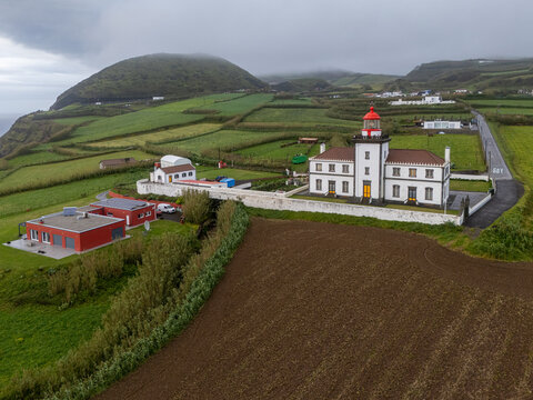 Aerial view of Farol da Ponta da Ferraria lighthouse surrounded by green fields, agricultural plots, and coastal cliffs in Ginetes, Azores, Portugal.