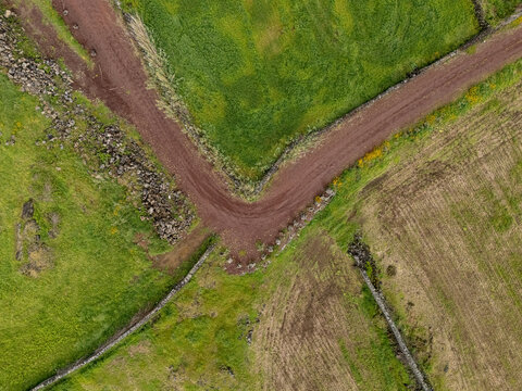 Aerial view of a dirt road intersection winding through lush green fields and traditional stone walls in the volcanic landscape of the Azores, Portugal.